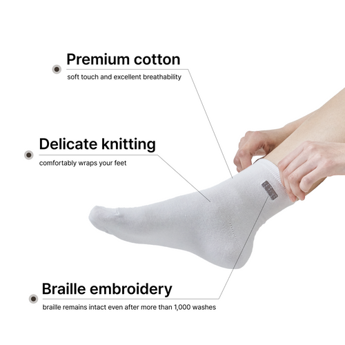 A person's leg and hands putting on a white premium cotton ankle sock featuring delicate knitting and a dark grey embroidered braille label